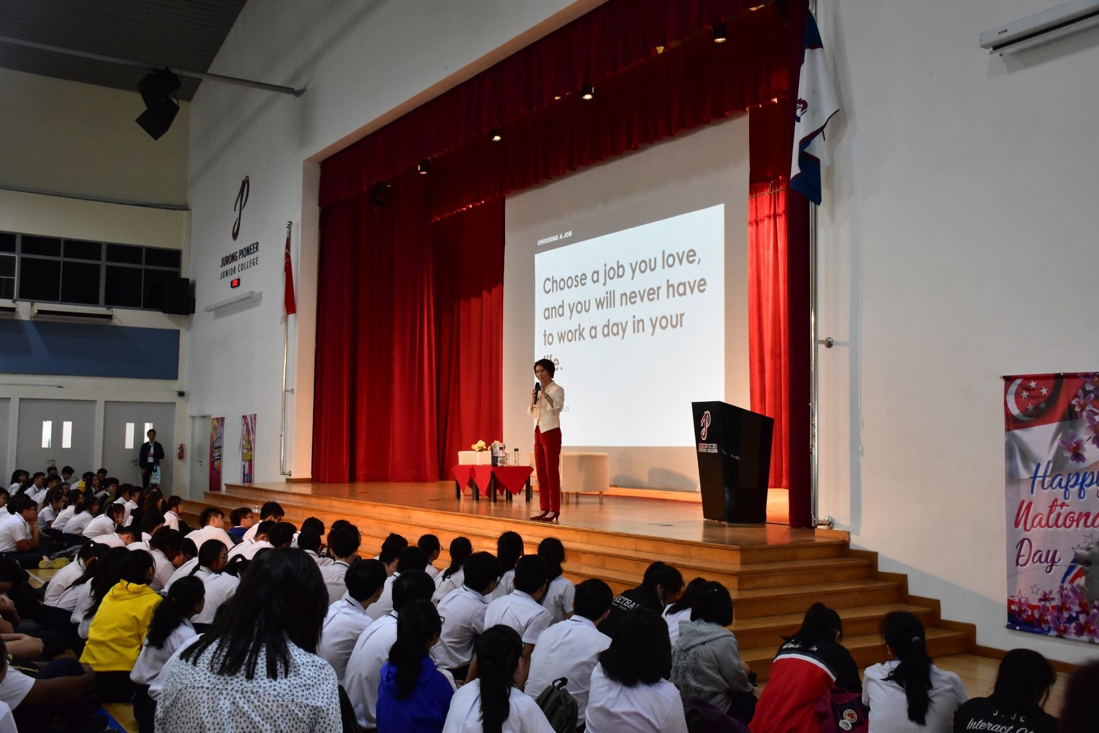 A woman speaks on stage to students in a school hall, with a slide reading "Choose a job you love" projected behind her.